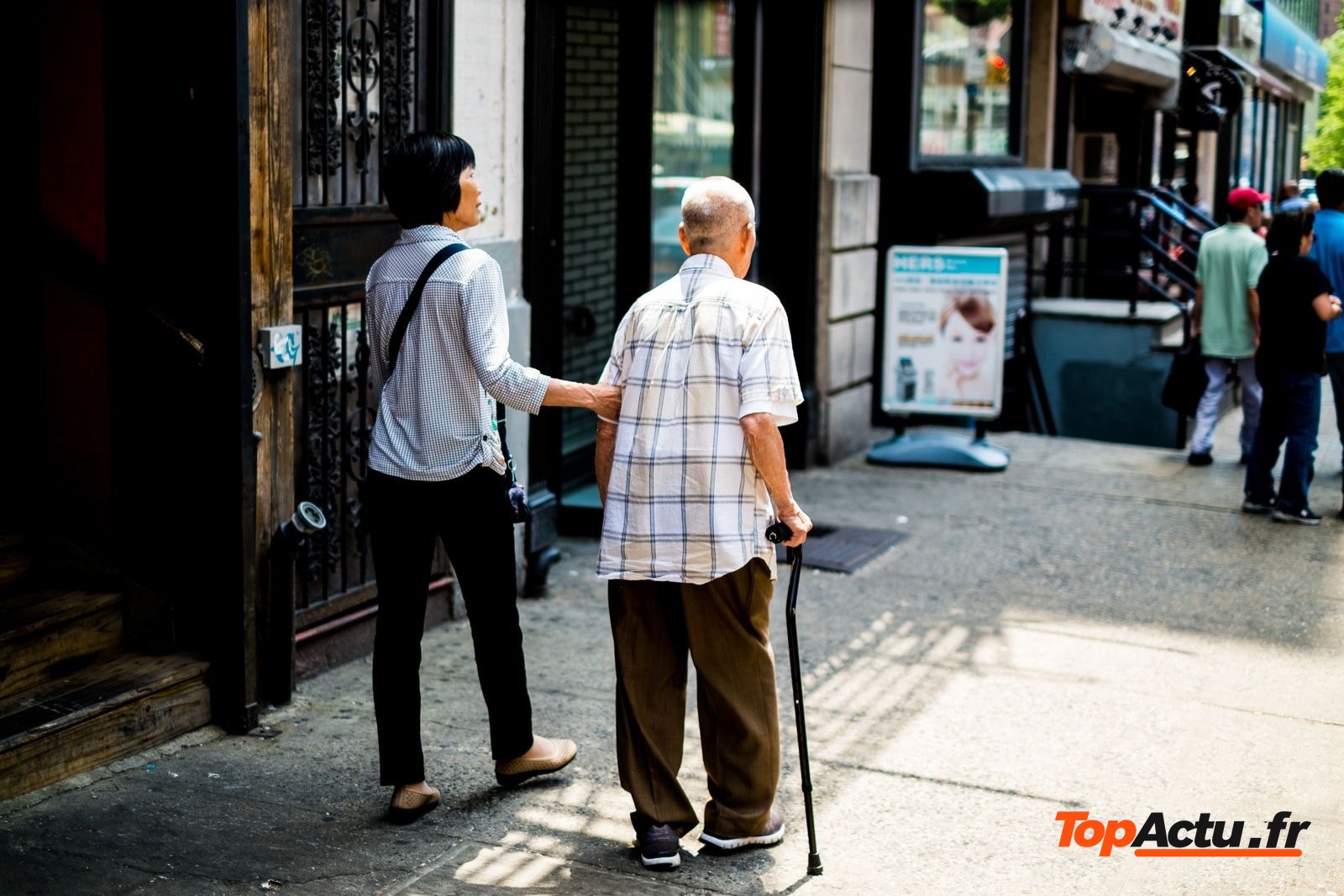 Une femme accompagne un homme âgé qui marche avec une canne dans une rue