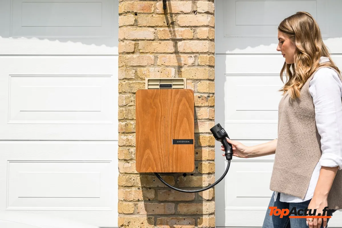 Une femme branche le câble d'une borne de recharge murale fixée sur un mur de briques, à côté d'un garage