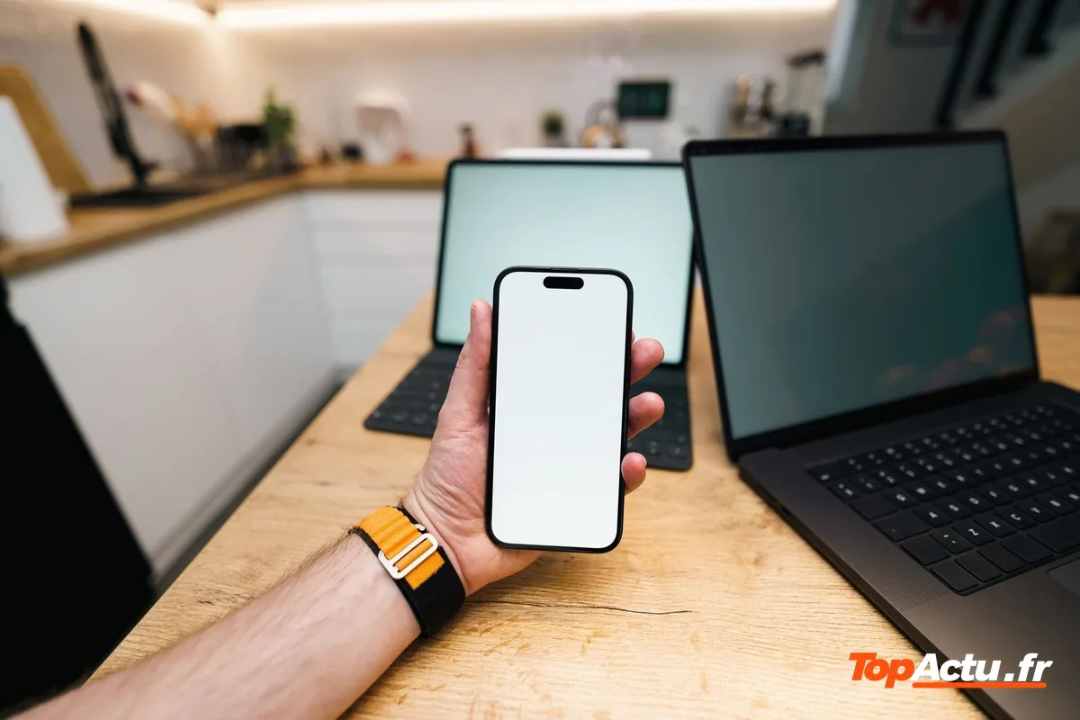 Une main qui tient un smartphone devant deux ordinateurs portables sur un plan de travail