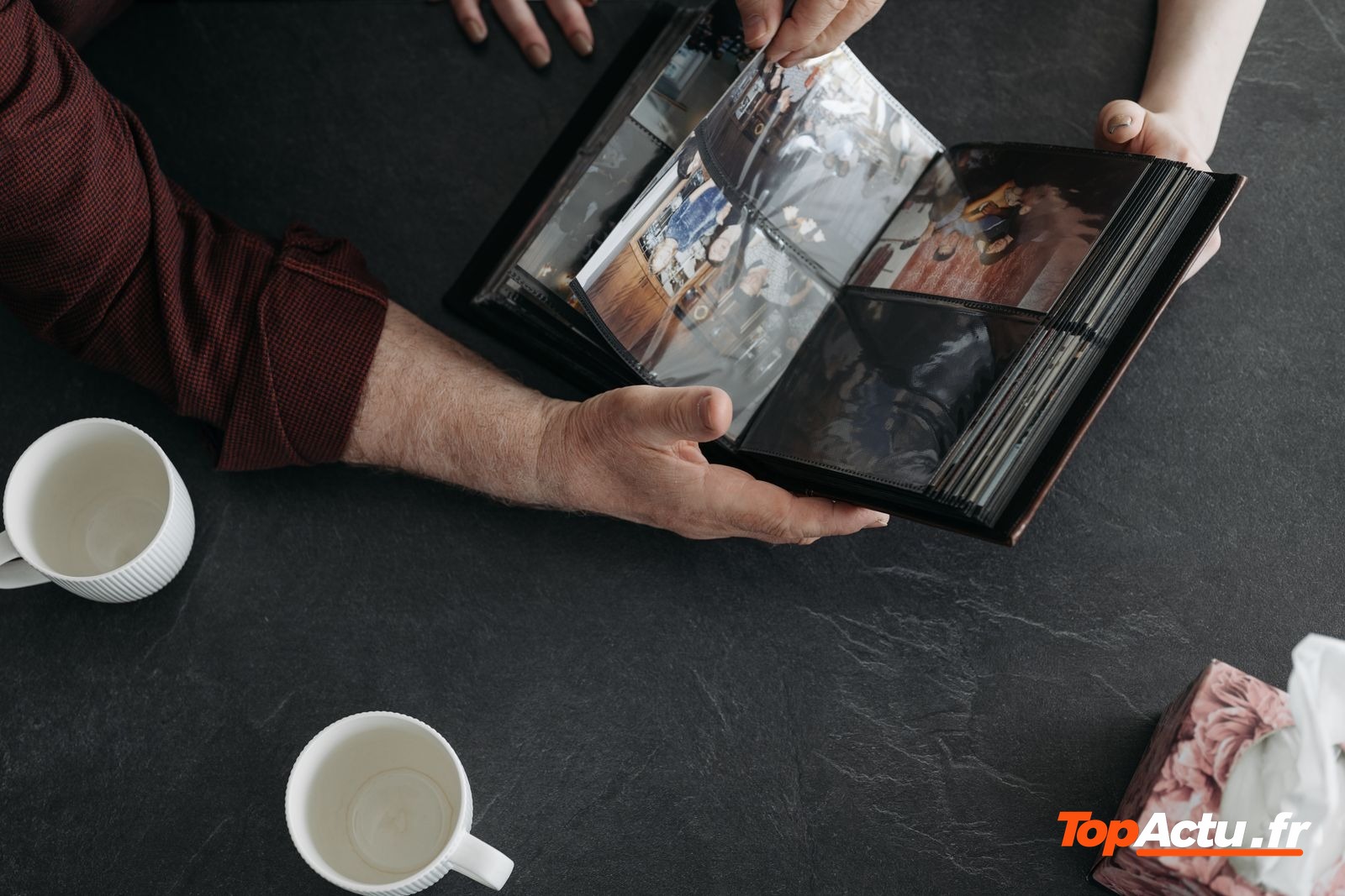 Mains qui tiennent un album photo ouvert, avec des tasses et une boîte de mouchoirs sur une table sombre
